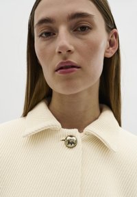 Krémový žebrovaný kabát s obráceným límcem a lesklým stříbrným knoflíkem, který má texturovanou látku a minimalistický design.