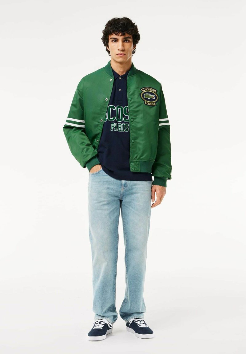Veste bomber en satin vert avec des rayures blanches, polo bleu marine avec le texte "LACOSTE PARIS", jeans bleu clair et baskets bleu marine.