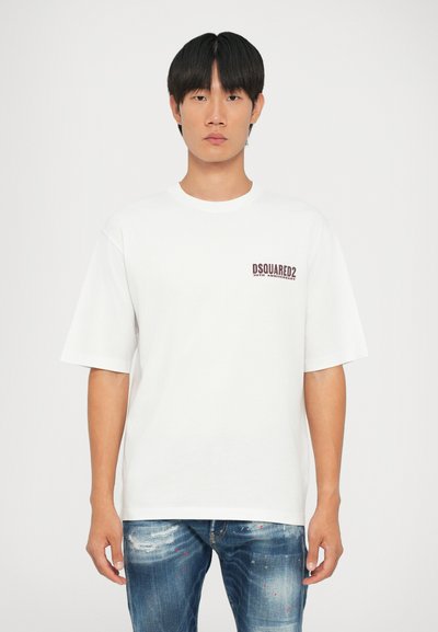 Camiseta blanca de algodón con cuello redondo y mangas cortas, que presenta un logotipo burdeos en el lado izquierdo del pecho. Combinada con jeans azules desgastados.
