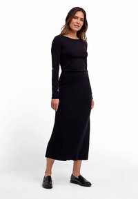 Schwarzes, geripptes Strickkleid mit langen Ärmeln, rundem Ausschnitt und tailliertem Schnitt, das bis zur Midi-Länge reicht, kombiniert mit schwarzen Schuhen.