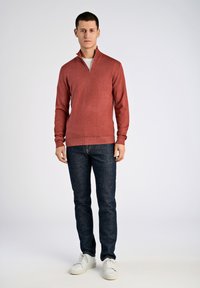 Sweat-shirt zippé rouge à manches longues, design ajusté, poignets côtelés, associé à un jean bleu foncé et des baskets blanches. Tricot simple et texturé.