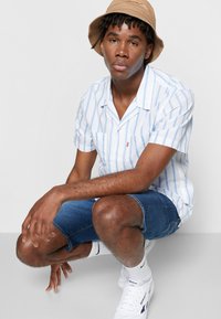 Camisa leve de mangas curtas, azul e branca às riscas, calções de ganga, sapatilhas brancas e um chapéu estilo balde bege.
