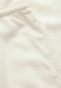 Tissu blanc avec une texture douce et lisse, doté d'une taille élastique et d'une poche latérale. Le matériau semble léger et respirant.