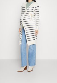 Stickad graviditetskofta med krämfärgad botten och marinblå ränder, inslagningsdesign, knytning i midjan, ihop med ljusblå utsvängda jeans och beige klackstövlar.