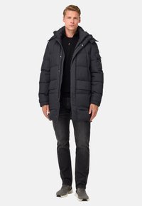 Man som står och har på sig en svart puffjacka med huva, svart tröja, mörka jeans och grå sneakers mot en enfärgad ljus bakgrund.