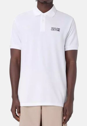 Mann trägt ein weißes Poloshirt mit kurzen Ärmeln und dem "Versace Jeans Couture" Logo auf der linken Brustseite sowie beigen Hosen.