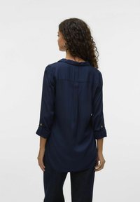 Blusa navy con colletto, maniche lunghe arrotolate e una leggera lucentezza. Presenta una piega sul retro e sottili dettagli di bottoni.