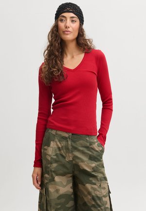 Femme aux cheveux bouclés portant un bandeau noir, un haut rouge à manches longues et un pantalon cargo camouflage vert, debout avec une main dans la poche.