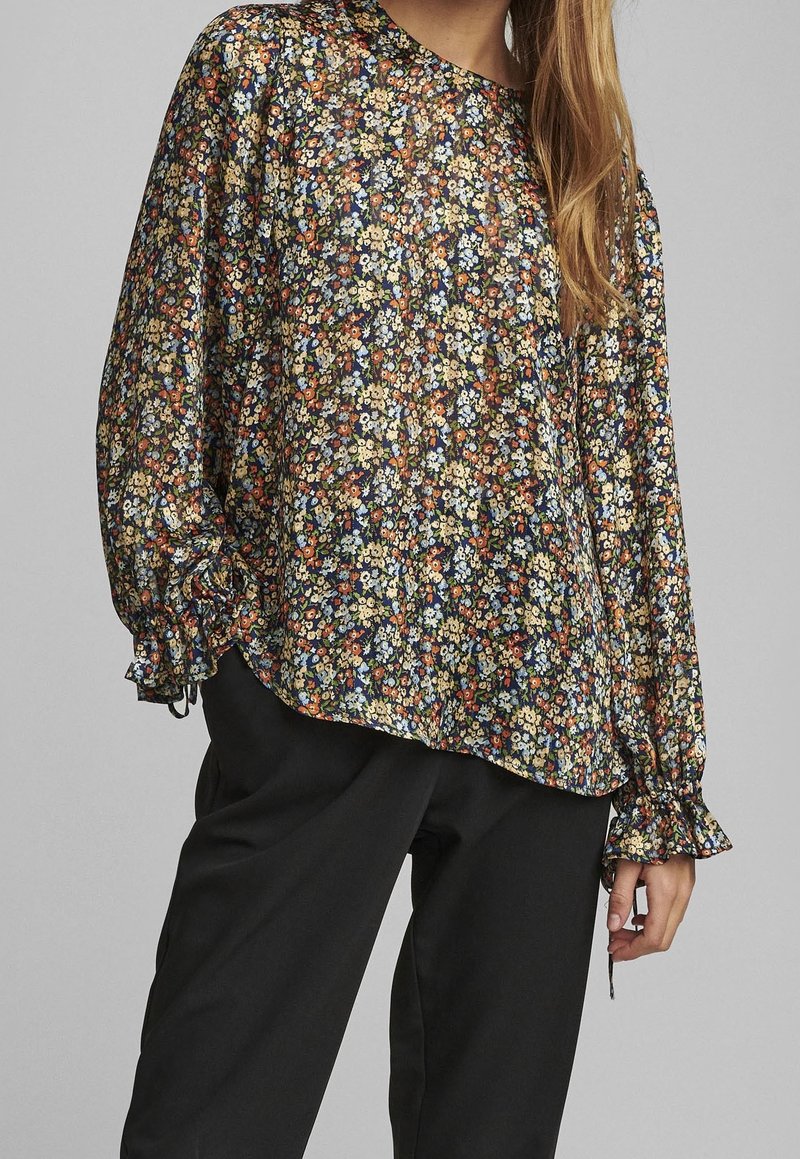Blusa floreale a maniche lunghe in tessuto scuro con sfondo nero. Presenta maniche a sbuffo con polsini elasticizzati e delicati motivi floreali in più colori.