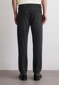 Widok z tyłu osoby w prostych czarnych dżinsach i czarnych butach stojącej przy białej strukturalnej ścianie i szarej podłodze.