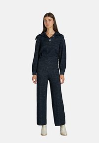 Kvinna iklädd en mörkblå ribbad stickad tröja med krage och matchande vida byxor, kompletterat med krämfärgade ankelskor, stående mot en enfärgad bakgrund.