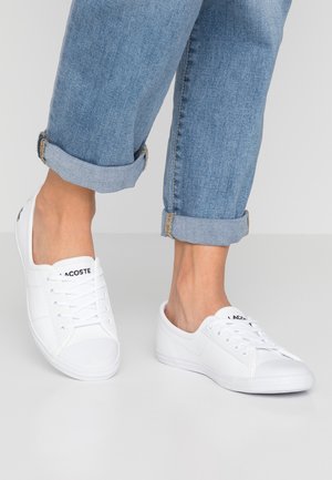 Osoba w niebieskich podwiniętych dżinsach i białych, niskich sneakersach Lacoste stoi na jasno szarej podłodze.
