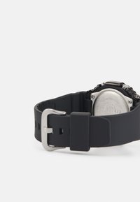 Bracelet de montre en silicone noir avec une boucle en métal argenté, présentant une surface texturée et un dos en acier inoxydable avec des détails gravés.