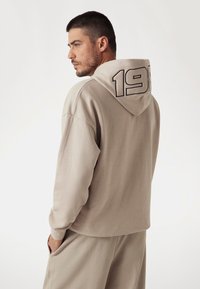 Beige színű kapucnis pulóver, húzózsinórral, amelyen egy nagy fekete "198" grafika található a hátán. Puha anyag és laza szabás jellemzi.