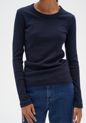 Frau trägt ein dunkelblaues, geripptes Langarmshirt und blaue Jeans, die Hände entspannt neben dem Körper vor einem einfarbigen Hintergrund.