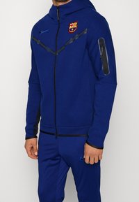 Felpa blu con zip realizzata in tessuto morbido, dotata di cappuccio, strisce nere diagonali e logo del Barcellona sul petto. Polsini e tasche texturizzati.