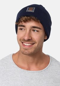 Laine de bonnette tricotée bleu marine avec un petit patch gris portant l'inscription "JEANSCODE". Le matériau semble doux et extensible, s'ajustant confortablement sur la tête.