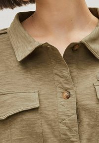Chemise olive verte avec un col boutonné, dotée de deux poches sur la poitrine, fabric texturé et détails de couture visibles.