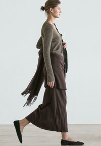 Brown knitted cardigan, brown long skirt, black flat shoes, and a fringe-accented scarf. Model is walking against a light background.