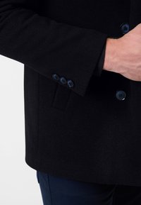 Zwarte wollen jas met een gladde textuur, voorzien van een double-breasted ontwerp en drie marineblauwe knopen op de manchetten, getoond met een hand.