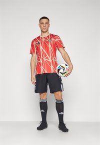 Camiseta de fútbol roja con patrones geométricos dorados, combinada con pantalones cortos negros y zapatillas negras. El jugador sostiene un balón de fútbol multicolor.
