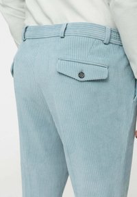 Hellblaue Cordhosen mit vertikalen Streifen, ausgestattet mit einer aufgesetzten Gesäßtasche mit Knopf und einem flachen Bund. Die Textur ist weich und gerippt.