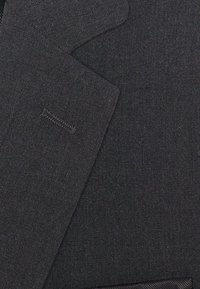 Donkergrijze stof met een gladde textuur. Heeft een reverskraag en subtiele stikseldetails, wat een klassiek design van een colbert benadrukt.
