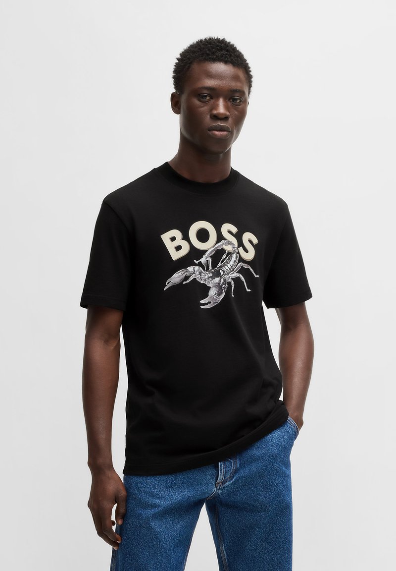 Juodas medvilninis marškinėliai su "BOSS" tekstu ir omaro grafika. Trumpomis rankovėmis, apvaliu kaklu ir laisvu siluetu, derinami su mėlynais džinsais.