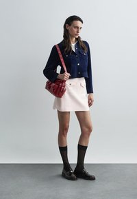 Veste courte en daim marine, top blanc, jupe rose, sac à main rouge, chaussettes noires montantes et mocassins en verni avec des accents dorés.