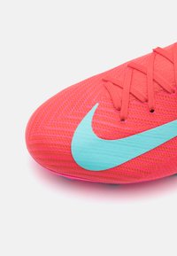 Jaskrawo różowe buty piłkarskie z teksturowaną powierzchnią, z dużym turkusowym logo Nike. Sznurowadła widoczne u góry, poprawiające chwyt.
