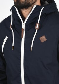 Marinejakke med hvit glidelås, lærdetaljer, justerbare snorer og teksturert finish med et logo-patch på brystet.
