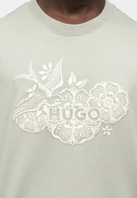 Gros plan d’une chemise vert clair avec un motif floral brodé blanc, des oiseaux et le texte « HUGO » sur la poitrine.