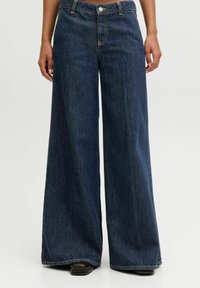 Person wearing dark blue wide-leg denim jeans, standing with hands relaxed at sides, shoes partially visible.