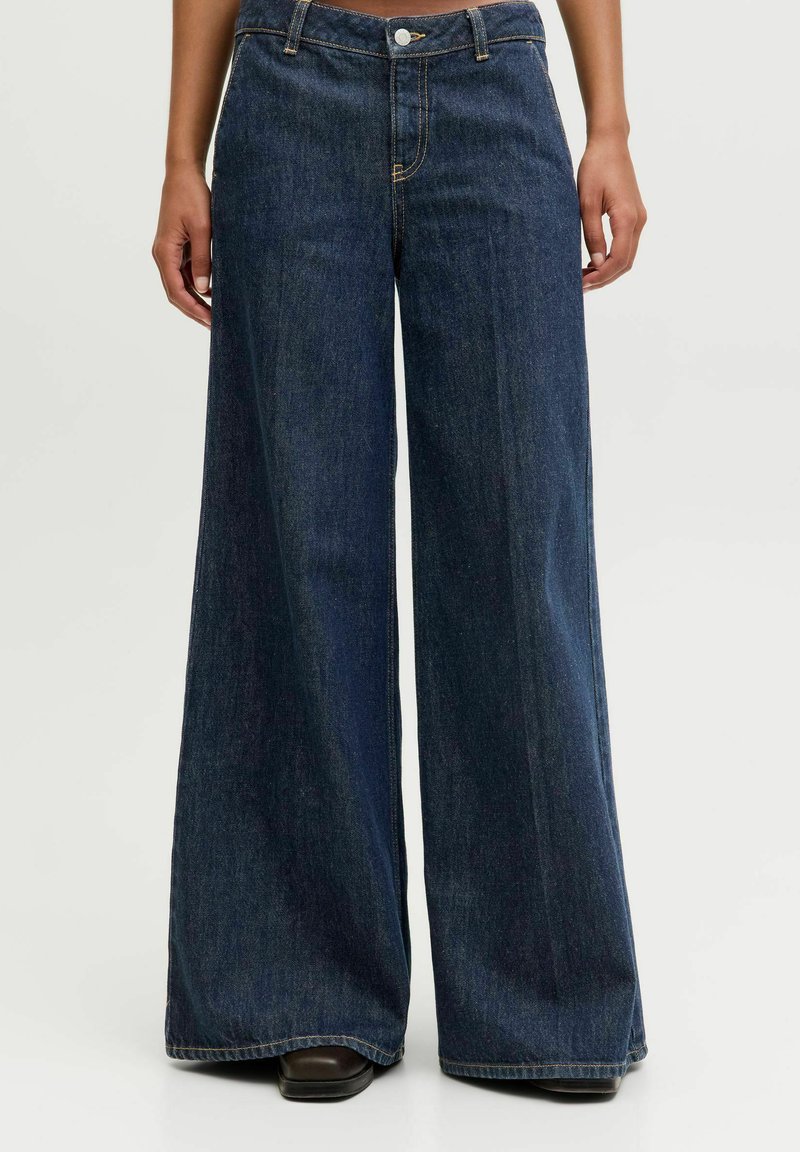 Person wearing dark blue wide-leg denim jeans, standing with hands relaxed at sides, shoes partially visible.