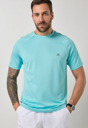 Uomo con barba e braccio tatuato che indossa una camicia a maniche corte azzurro chiaro e pantaloni bianchi, in piedi con le mani nelle tasche.