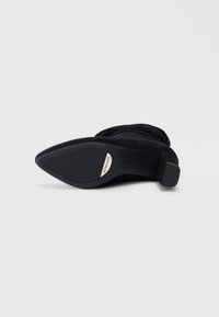 Botte en suède noire à talons hauts avec un bout pointu, texture lisse, talon effilé et détail de logo métallique sur la semelle.