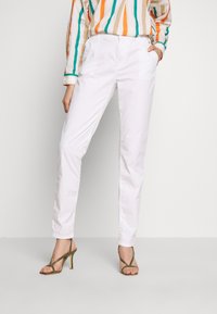 Femme portant un pantalon blanc ajusté, une chemise à rayures à manches longues, et des sandales à talons hauts vert olive, debout avec une main dans la poche.