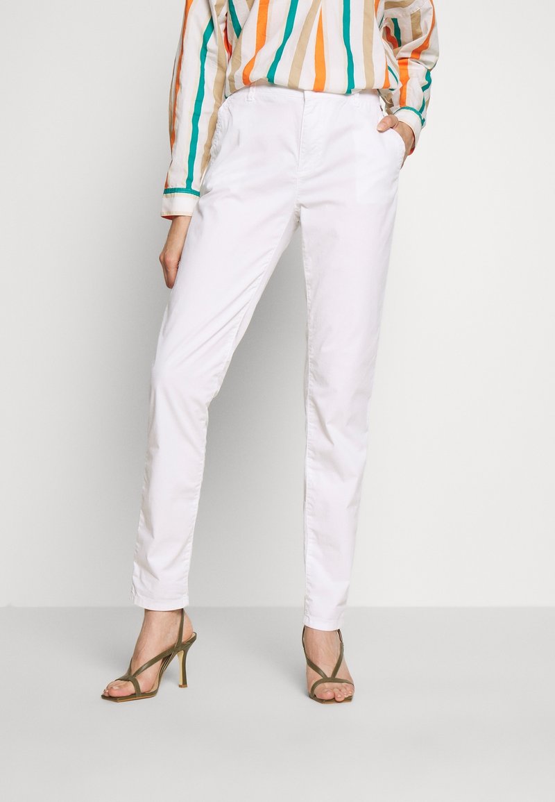 Femme portant un pantalon blanc ajusté, une chemise à rayures à manches longues, et des sandales à talons hauts vert olive, debout avec une main dans la poche.