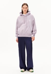 Lavenderfarbener Kapuzenpullover mit einer vorderen Tasche und einem weißen Außenlogo, kombiniert mit navyblauen, weit geschnittenen Hosen und weißen Sneakers mit grünen Akzenten.