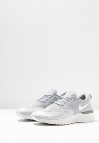 Zapatillas deportivas gris claro con una parte superior de malla transpirable, cuello acolchado y suela de goma blanca con un diseño texturizado y el logo de Nike.