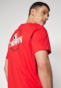 Rød bomulds T-shirt med korte ærmer, der har et stort rundt Champion-logo og tekst på ryggen. Glat tekstur, rund hals.