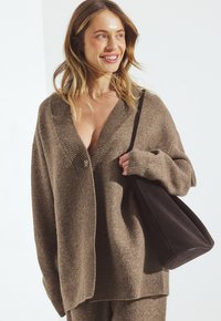 Brūns pārāk liels kardigans ar dziļu V-veida kaklu, ribotām piedurknēm un vienu pogu. Valkāts ar melnu plecu somu.