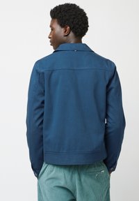 Marineblauwe denimjas met een klassieke kraag, knoopmanchetten en een rechte pasvorm. Platte naden en een subtiele rugpasdetail.