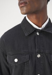 Zwarte denimjas met een klassieke kraag, zilveren knopen en twee borstzakken. De stof heeft een gladde textuur en een gestructureerd ontwerp.
