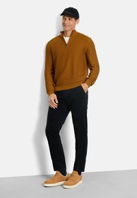 Brauner Zip-Pullover aus strukturiertem Stoff, kombiniert mit schwarzen Cordhosen und beigefarbenen Turnschuhen. Das Model trägt außerdem eine schwarze Mütze.