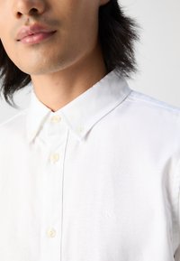 White button-up shirt with a spread collar, featuring textured fabric, cream buttons, and a subtle embroidered logo on the chest.