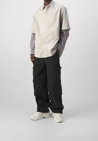 Mężczyzna w warstwowej, długiej koszuli w kratę noszonej pod koszulą z krótkim rękawem w paski, czarnych spodniach cargo i białych, masywnych sneakersach.