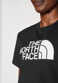 Melnas kokvilnas t-krekls ar lieliem baltiem burtiem "THE NORTH FACE" un izliekta logotipa dizainu priekšpusē. Īsas piedurknes un apļa kakls.