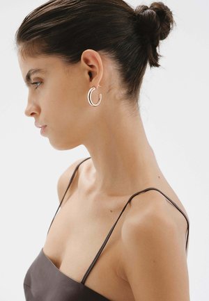 Pendiente de aro en metal de oro rosa, con un diseño elegante y minimalista que presenta un sutil detalle de bisagra. Mostrado en la oreja de un modelo.