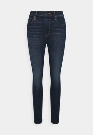 Jean slim bleu foncé avec cinq poches, fermeture à bouton et glissière à l'avant, présenté sur un fond blanc uni.